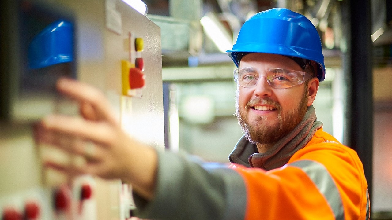 Een elektrotechnisch ingenieur lacht naar de camera terwijl hij op een knop op een bedieningspaneel drukt.