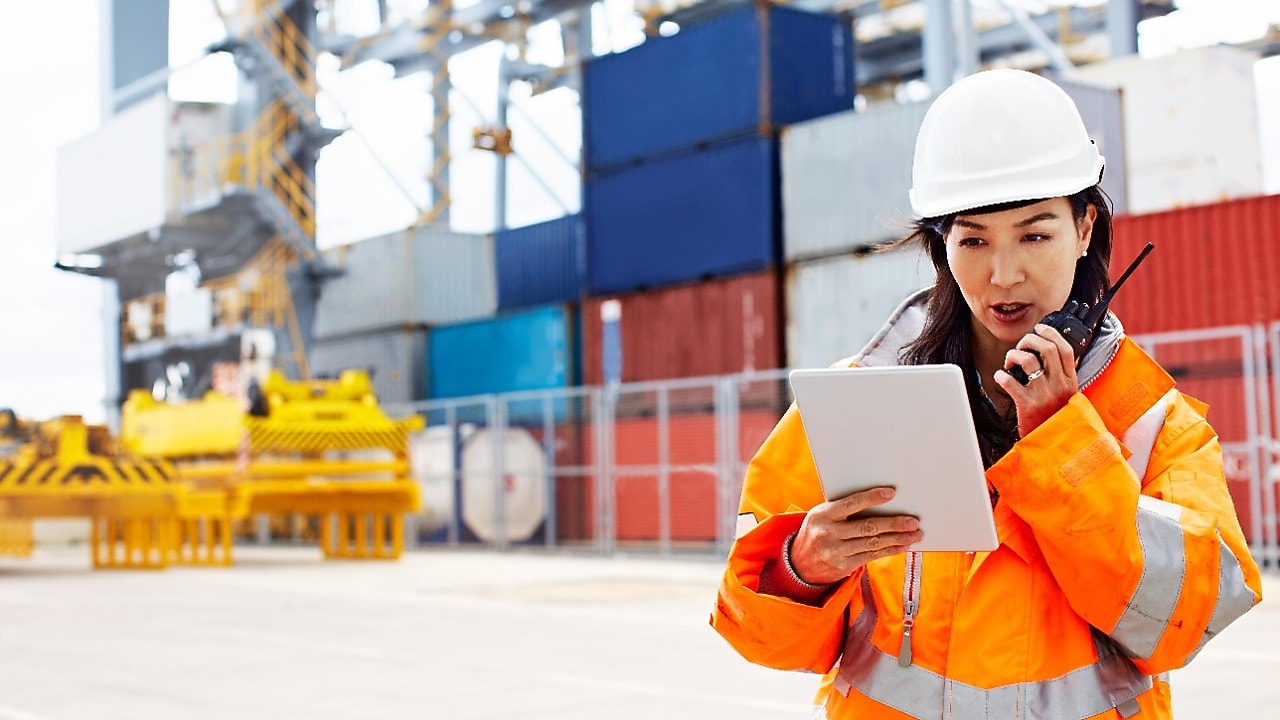 Een medewerkster kjkt op een tablet en spreekt in een walk-talkie, op de achtergrond opgestapelde zeecontainers