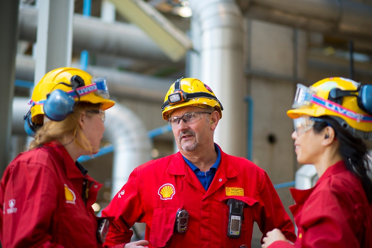 Drie medewerkers in rode overall en gele valhelm op praten met elkaar. Shell Energy and Chemicals Park Rotterdam (Pernis)