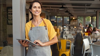 Portrait of a happy waitress standing at restaurant entrance holding digital tablet.