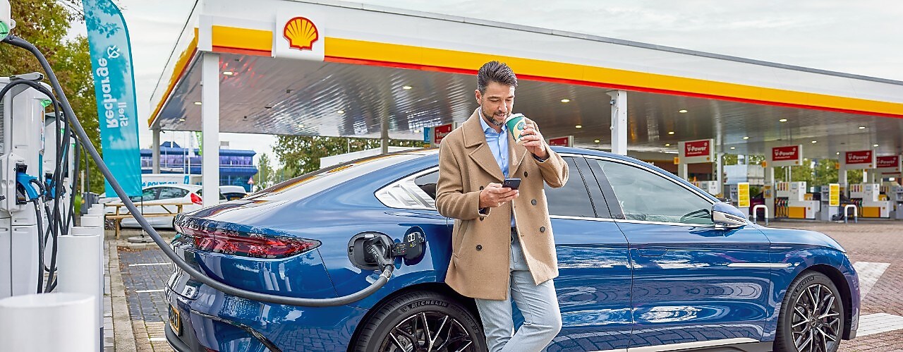 Een man kijkt op zijn mobiel en drinkt koffie terwijl zijn blauwe elektrische auto aan de snellader staat bij een Shell-retailstation