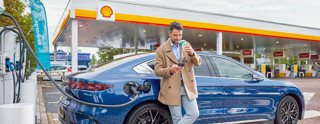 Een man kijkt op zijn mobiel en drinkt koffie terwijl zijn blauwe elektrische auto aan de snellader staat bij een Shell-retailstation