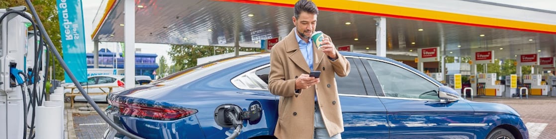 Een man kijkt op zijn mobiel en drinkt koffie terwijl zijn blauwe elektrische auto aan de snellader staat bij een Shell-retailstation