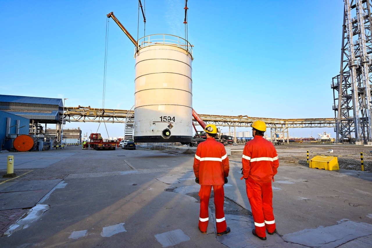Twee medewerkers in rode overall en met gele bescherminghelm kijken hoe de opslagtank van zijn plaats wordt gehesen (Foto: Ernst Bode)