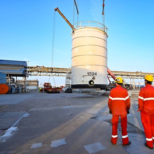 Twee medewerkers in rode overall en met gele bescherminghelm kijken hoe de opslagtank van zijn plaats wordt gehesen (Foto: Ernst Bode)