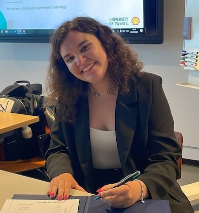 The photo shows Valeria (head and shoulders), with shoulder-long curly hair wearing a black blaser on a white top. She sits at a desk in a class room or conference room and smiles towards the camera. Behind her the down edge of a smart board with a Shell presentation is visible. (Courtesy of Valeria Pinus)