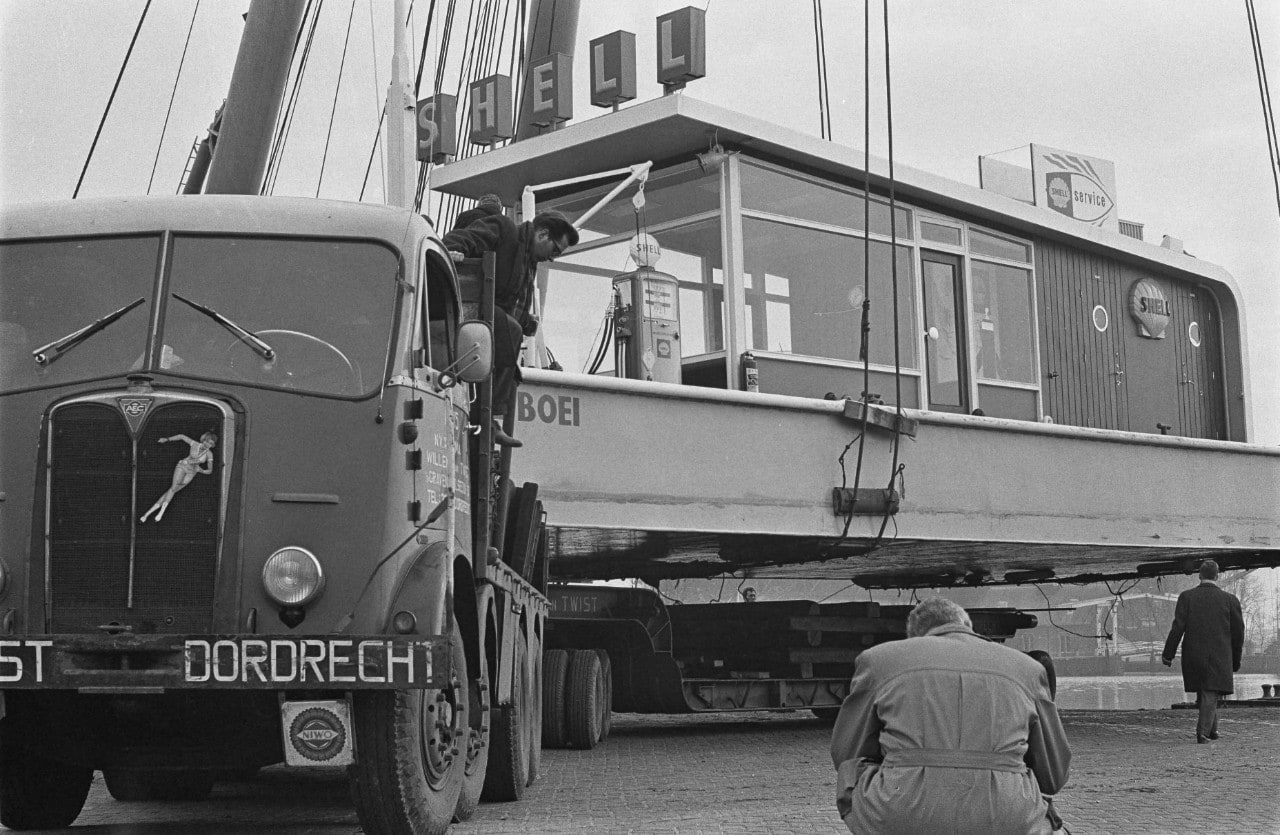 Op de foto wordt het station — „Shell Boei" geheten na uit de Amstel te zijn gehesen, op wielstellen naar het nieuwe RAI-gebouw in Amsterdam getransporteerd, waar het op de tentoonstelling Hiswa - Goed Kamp zou komen te staan, 14 maart 1963 (Foto: Hugo van Gelderen/Anefo/Nationaal Archief)