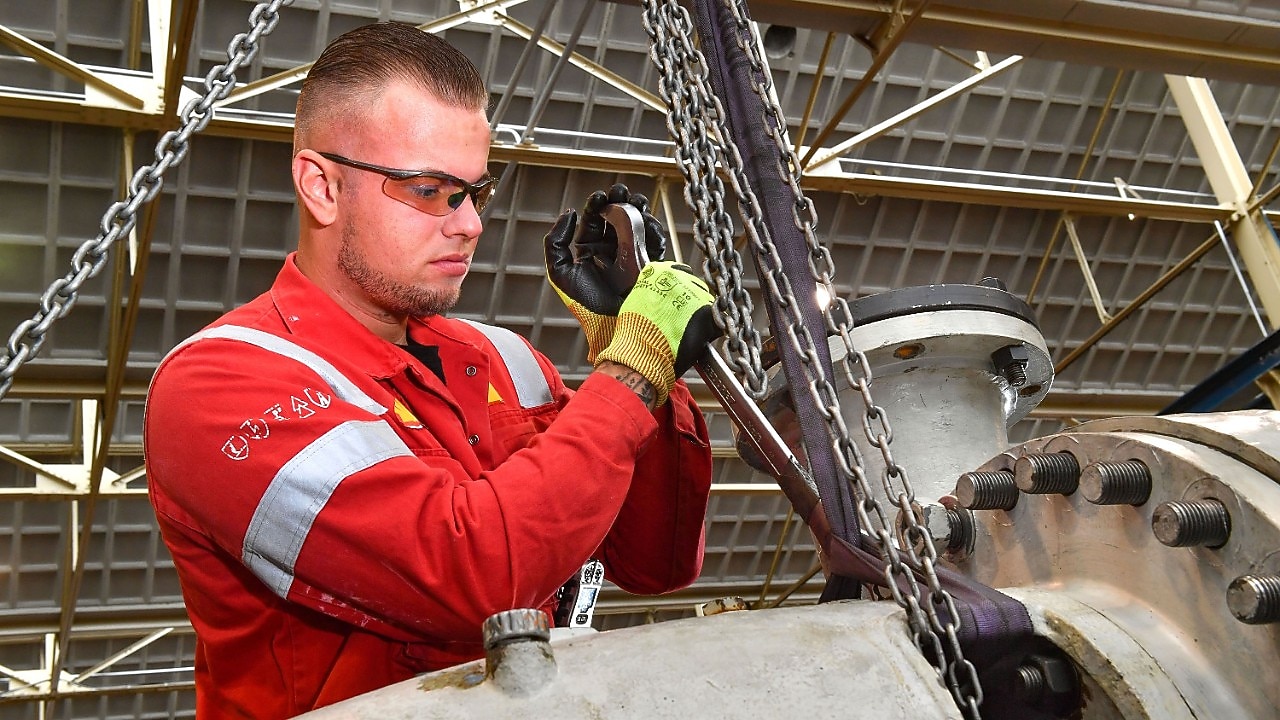 Stefan de Visser in de weer in de Centrale Werkplaats van Shell Pernis. Hij heeft kort haar, draagt een beschermbril en handschoenen. Hij heeft een grote sleutel van ongeveer een 50 centimeter groot vast waarmee hij iets aandraait op een metalen constructie (Beeld: Ernst Bode)