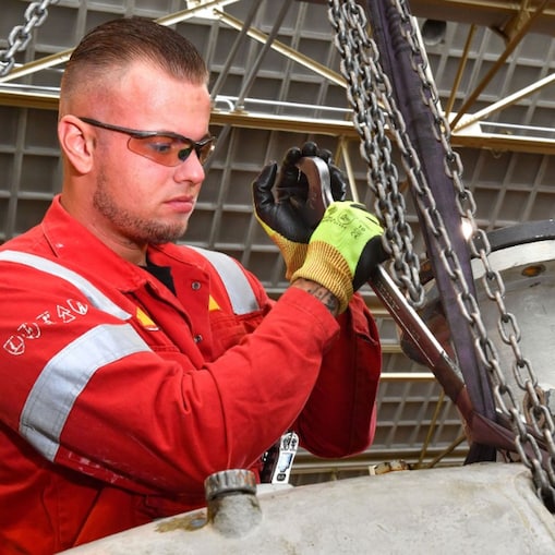 Stefan de Visser in de weer in de Centrale Werkplaats van Shell Pernis. Hij heeft kort haar, draagt een beschermbril en handschoenen. Hij heeft een grote sleutel van ongeveer een 50 centimeter groot vast waarmee hij iets aandraait op een metalen constructie (Beeld: Ernst Bode)