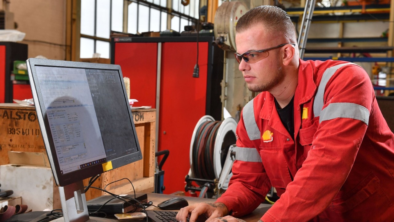 Stefan bekijkt een werkorder op een computerscherm. Hij heeft zijn handen op het toetsenbord. Op de achtergrond is de werkplaats zichtbaar (Beeld: Ernst Bode)