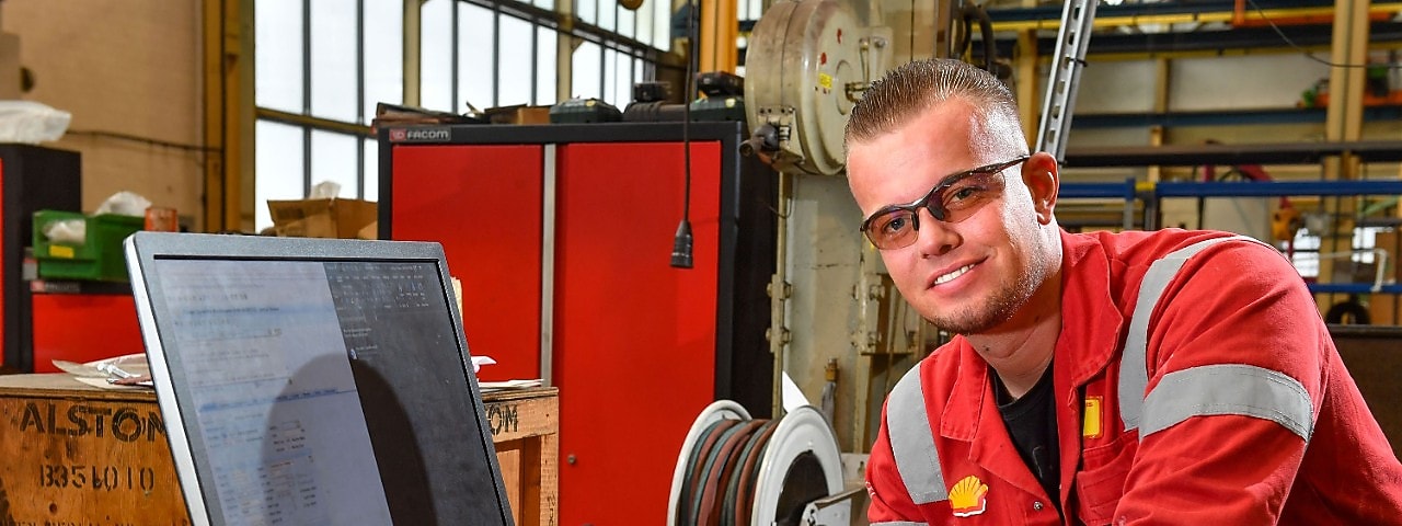 Stefan smiles towards the viewer. He has short hair, wears safety glasses and a red protective overall. A computer screen with a work order is close to him. In the background parts of the Central Workshop of Shell Pernis are visible (Photo: Ernst Bode)