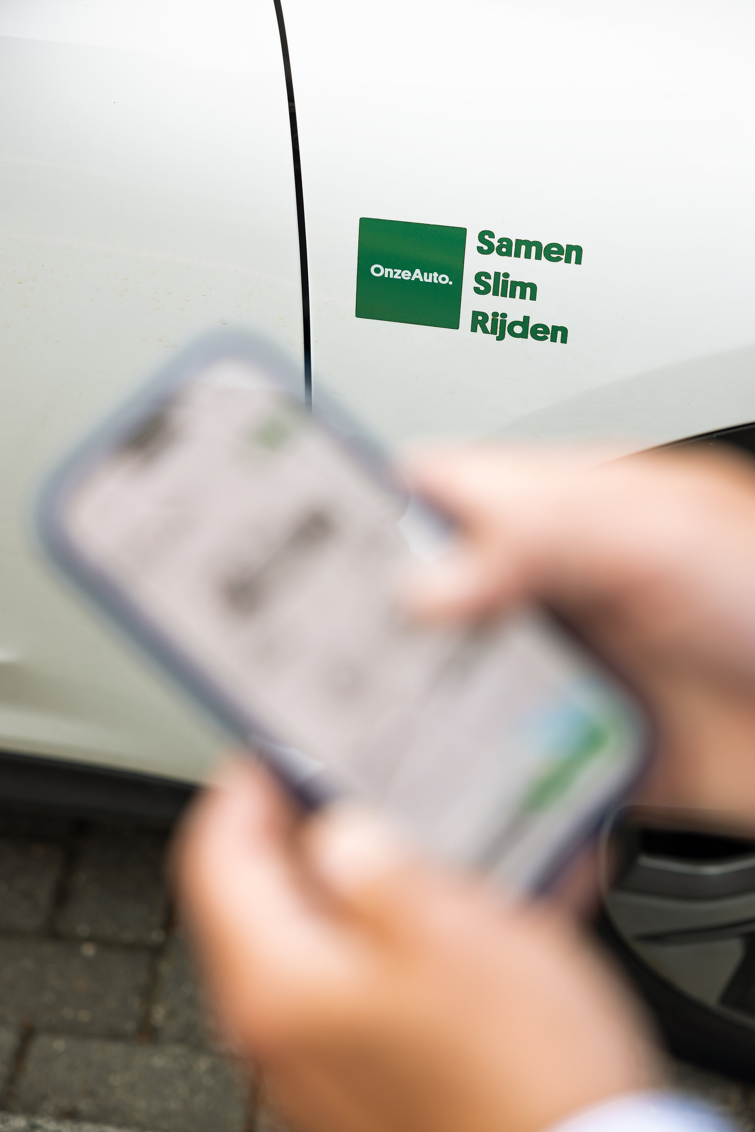 De app van OnzeAuto op de mobiel met op de achtergrond het logo van OnzeAuto. Samen Slim Rijden. (Foto: Christian Kalse)