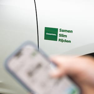 De app van OnzeAuto op de mobiel met op de achtergrond het logo van OnzeAuto. Samen Slim Rijden. (Foto: Christian Kalse)
