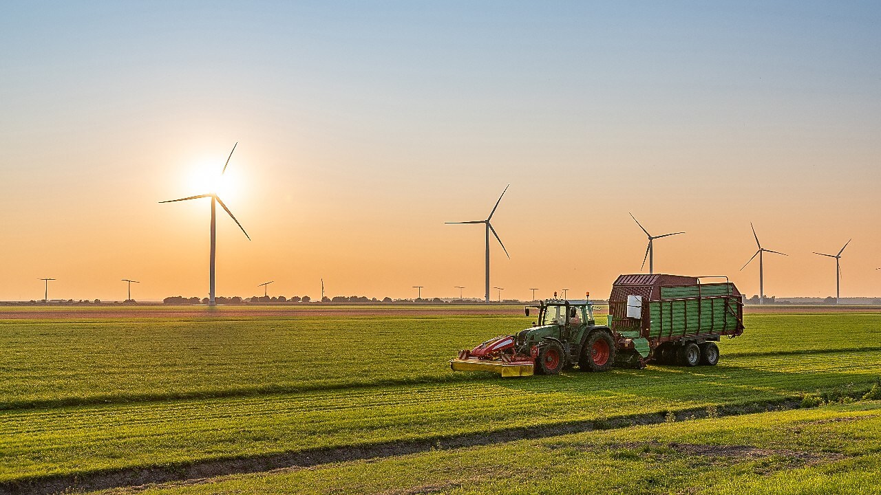 Een tractor met trailer op het veld, met op de achtergrond windturbines en een opkomende zon