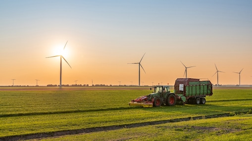 Een tractor met trailer op het veld, met op de achtergrond windturbines en een opkomende zon