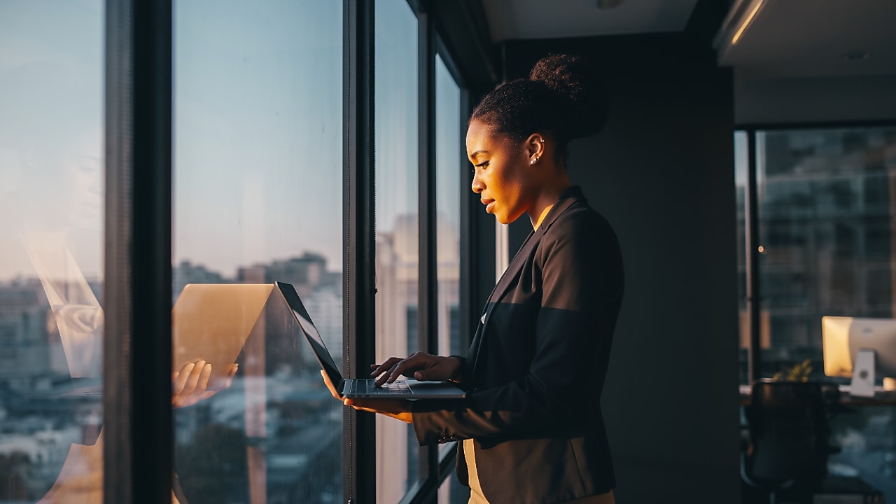 Een zakenvrouw kijkt op haar laptop, staande bij het raam met uitzicht over de stad (Foto: AI-gegenereerd met Adobe Firefly)