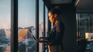 Een zakenvrouw kijkt op haar laptop, staande bij het raam met uitzicht over de stad (Foto: AI-gegenereerd met Adobe Firefly)