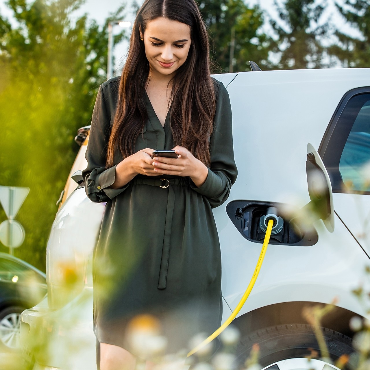 Een vrouw kijkt op haar mobiel terwijl ze haar witte elektrische auto laadt