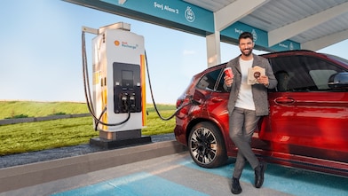 Een man met koffie en snack terwijl zijn auto oplaadt bij een Shell Recharge-lader onderweg.